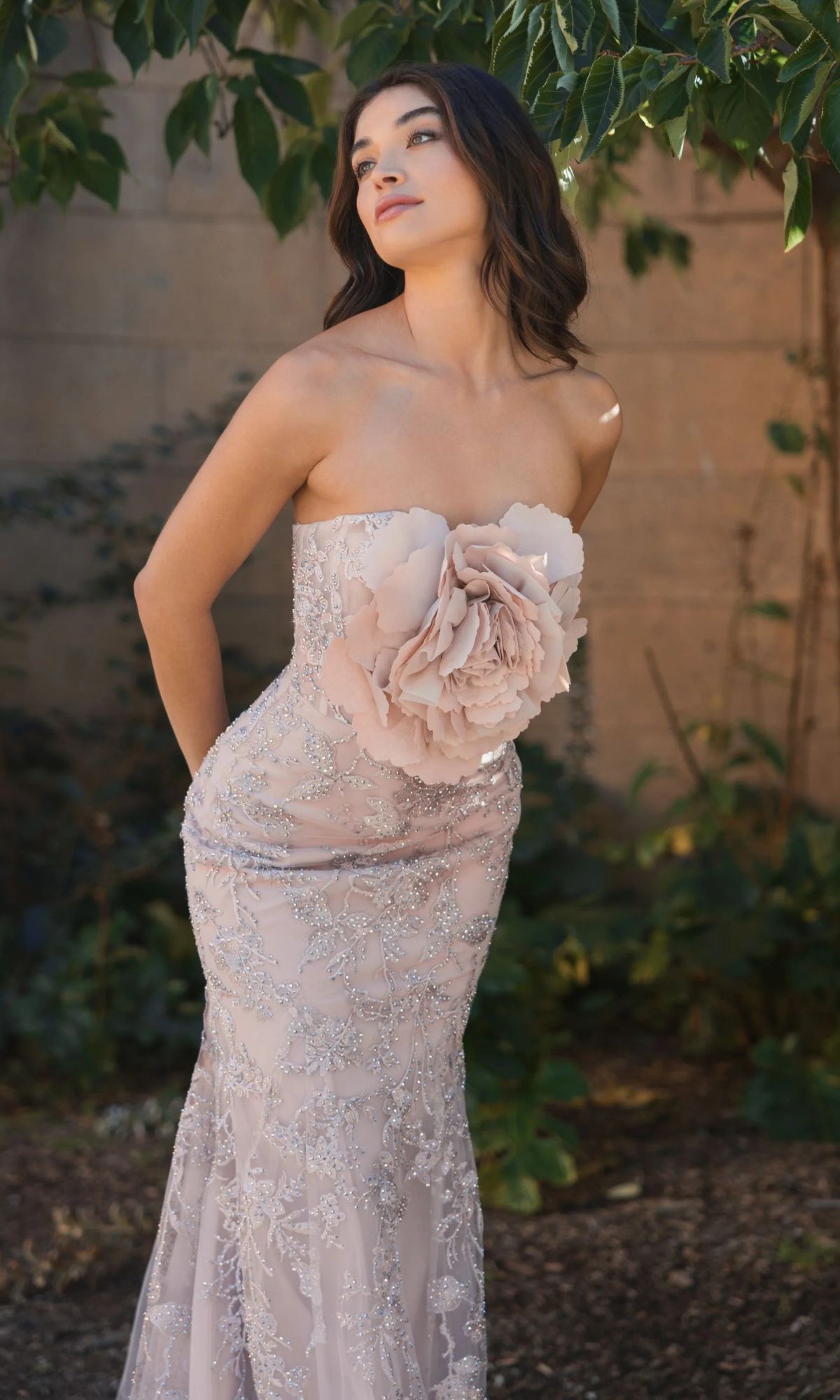 A woman wears the Mac Duggal 21036 Strapless Long Floral Prom Dress, featuring silver embellishments and a large floral waist detail, as she stands outdoors beneath tree branches.
